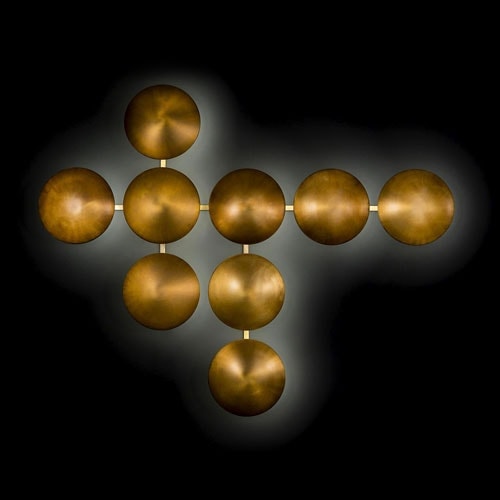 Wall Clips LED lamp made up of round burnished bronze steel modules joined by a golden metal rod on a black wall.