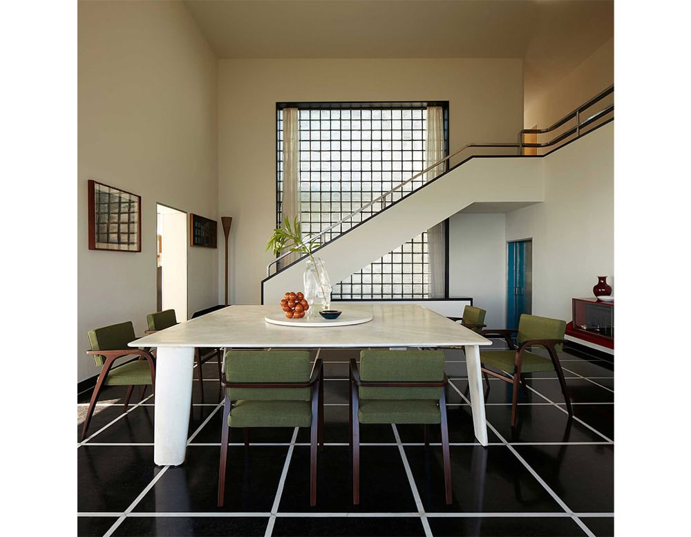 A Biscuit Dinner Table with a light-colored surface and thick, rectangular legs is shown in a modern dining room; the table is surrounded by four green chairs, with a staircase and a large window in the background