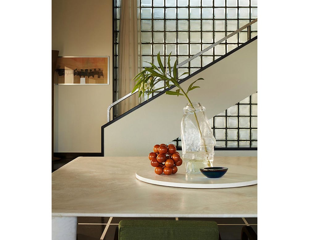 A light wood Biscuit Dinner Table is decorated with a centerpiece with red spheres and a vase. A staircase with a geometric railing is visible in the background.