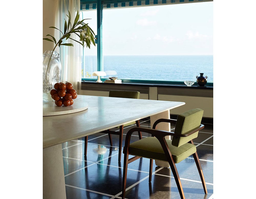 A light-colored Biscuit Dinner Table with a green chair is shown in a dining area with an ocean view. A vase with greenery and red decor are on the table.