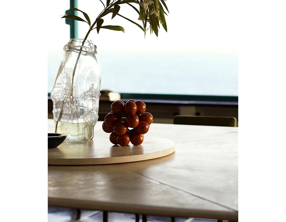 A Biscuit Dinner Table with a light-colored surface is shown with a glass vase holding greenery, red spheres on a tray, and a dark object. A window with a view is in the background.