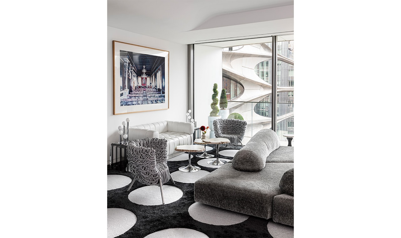 A contemporary living space featuring a gray modular sofa, patterned black-and-white rug, and accent chairs, with floor-to-ceiling windows showcasing an architectural view