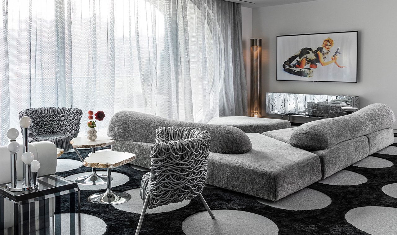 A modern living area with a plush gray lounge sofa, a uniquely designed accent chair, and a patterned black-and-white rug, complemented by soft sheer curtains filtering natural light