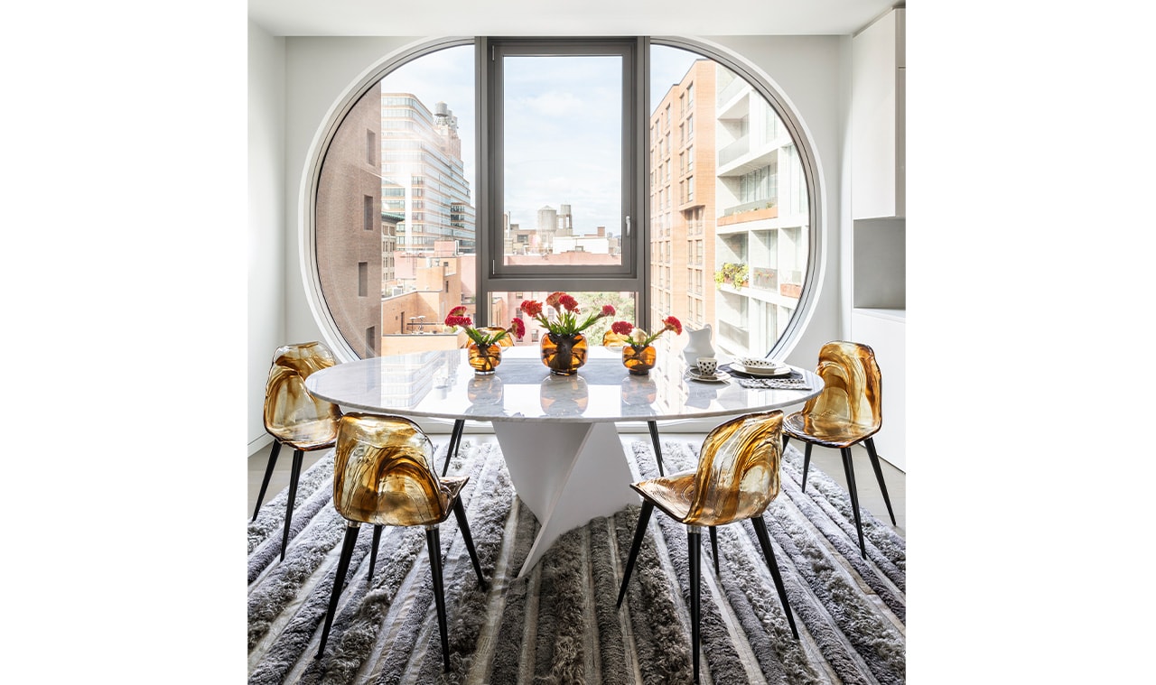A contemporary dining space with a round window framing a city view, featuring a sleek white table, gold-accented chairs, and a patterned rug for a sophisticated ambiance
