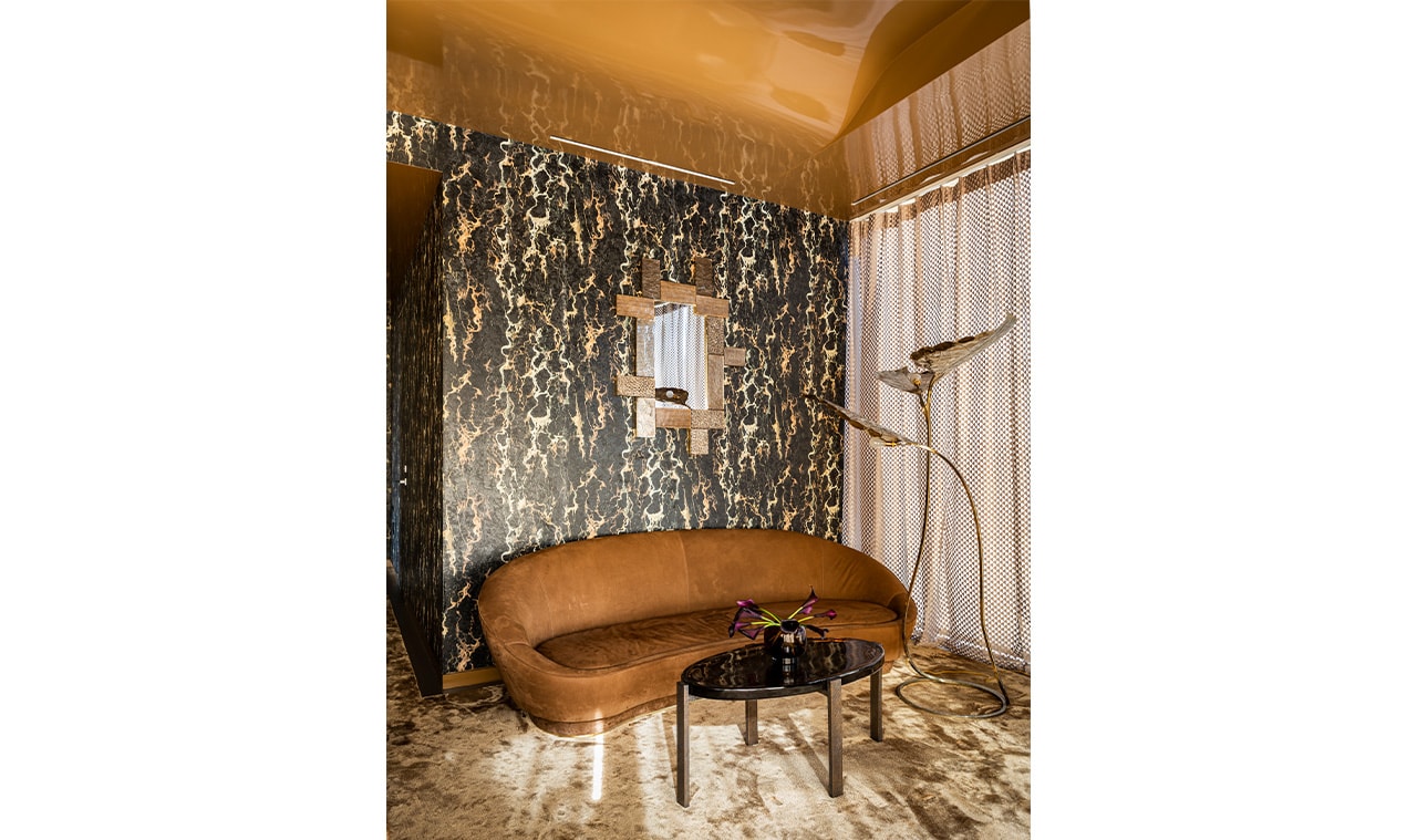 A luxurious lounge area with a curved brown sofa, a round black coffee table, and gold-accented decor against a dramatic patterned wall and warm lighting