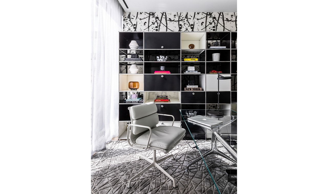 A contemporary home office with a black modular bookshelf, two modern leather chairs, a glass coffee table, and a plush textured rug under natural light