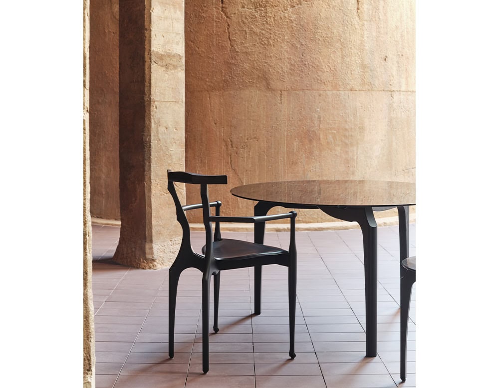 BD Carlina Round Dining Table with a dark, sleek finish and slender legs, paired with a minimal black chair featuring curved lines. The scene is framed by warm stone walls and tiled flooring, adding texture and depth.