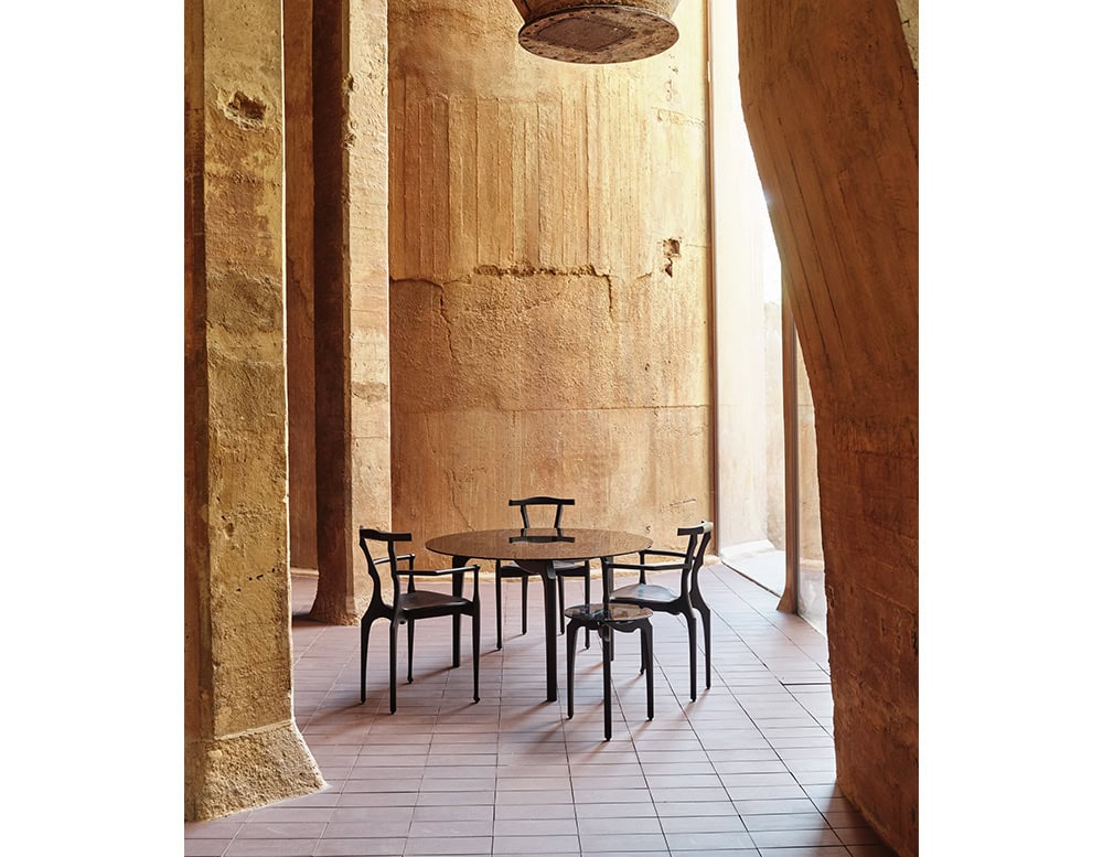BD Carlina Round Dining Table crafted with a sleek dark finish and thin, elegant legs for a light, architectural look. Four minimal black chairs surround it. The scene is set in a warm space with textured stone walls and tiled floors.