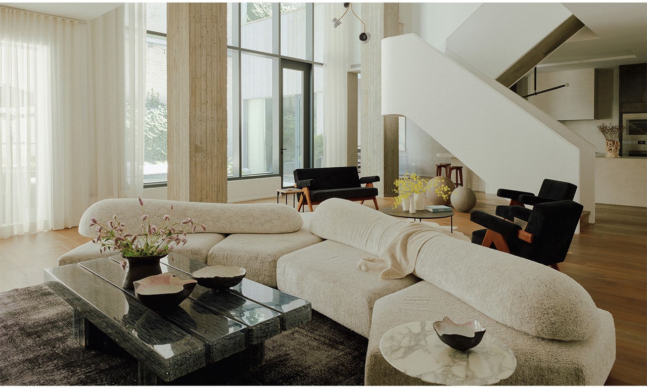 A sophisticated living space featuring a grand white staircase, plush cream seating, and dark marble coffee tables, bathed in natural light from floor-to-ceiling windows