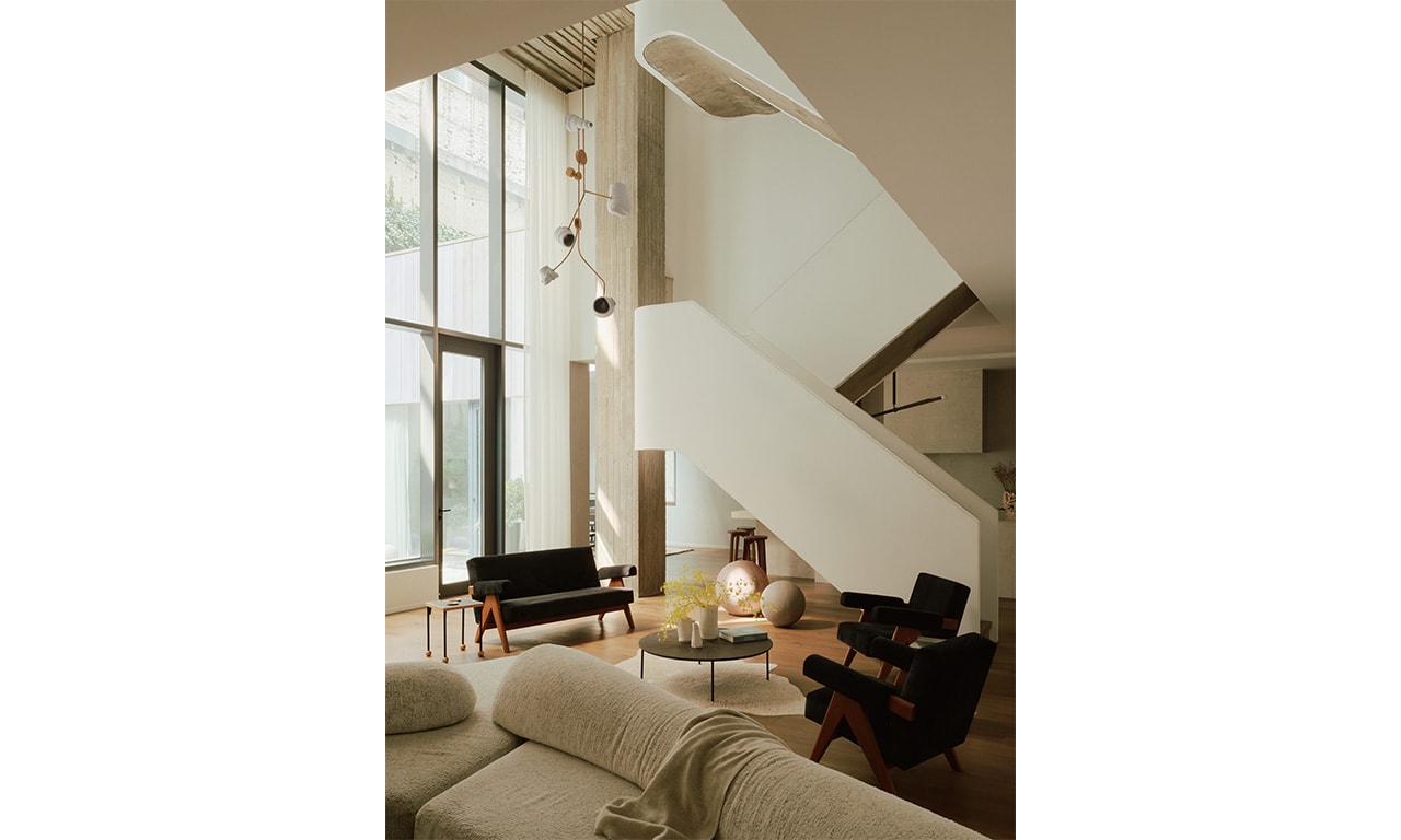 A spacious living area with high ceilings and a sculptural staircase, featuring modern furnishings and floor-to-ceiling windows that enhance natural light