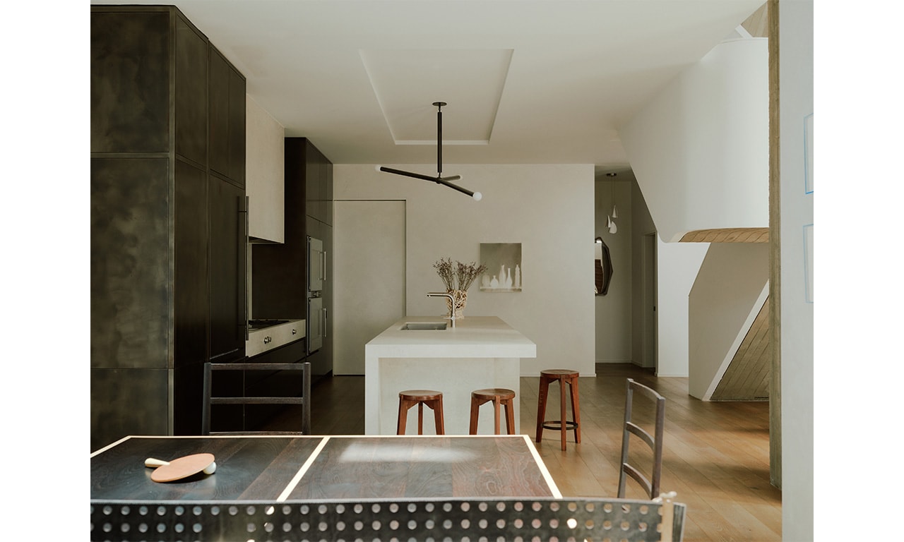 A modern kitchen featuring a sleek white island, wooden stools, and minimalist cabinetry, complemented by a black accent wall and a sculptural light fixture