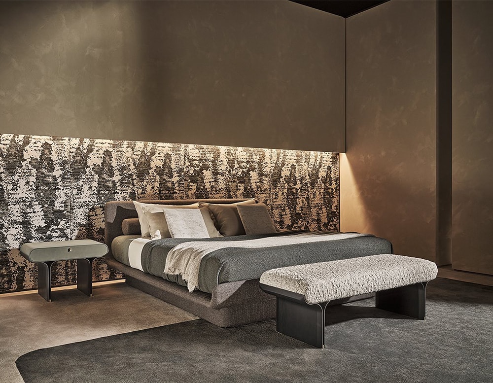 Bench with curved black stained ash base and bright brass details with a cushion covered by clear grey fabric in a room near to a bed