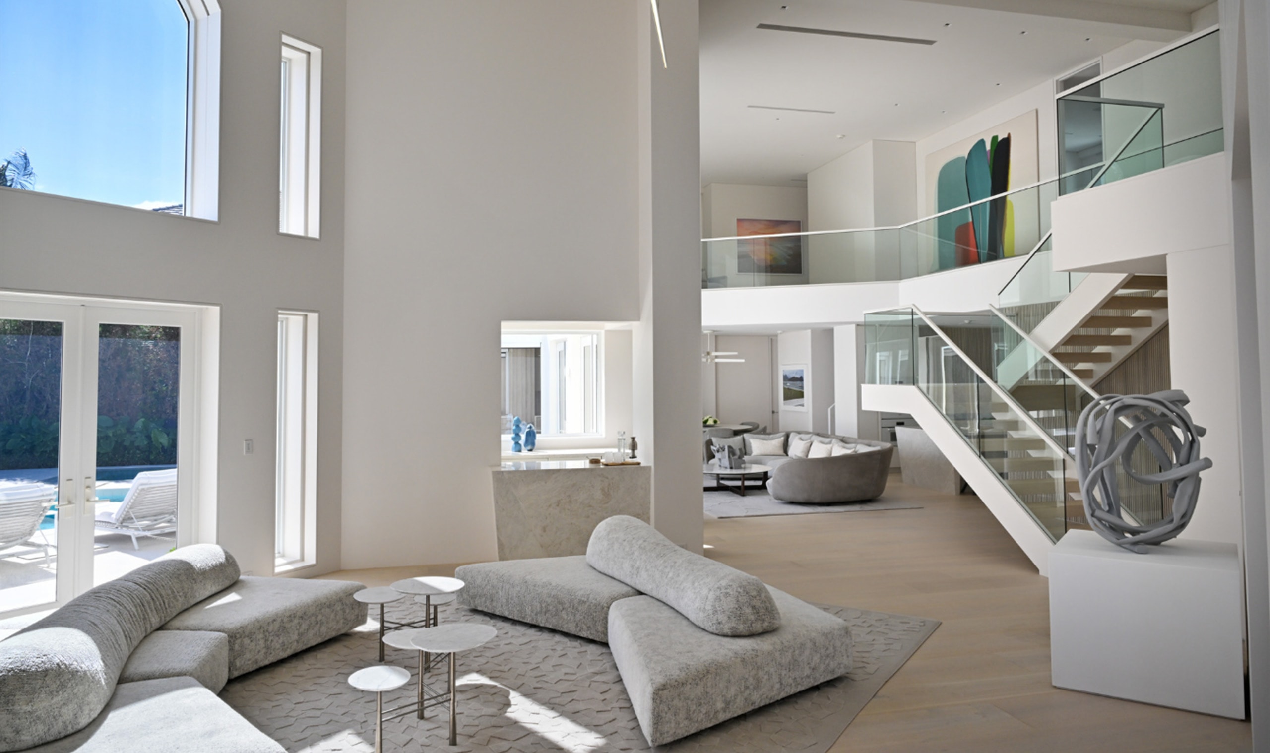 An airy open-concept living space featuring sleek modern furnishings, towering windows, and a sculptural staircase for a refined aesthetic