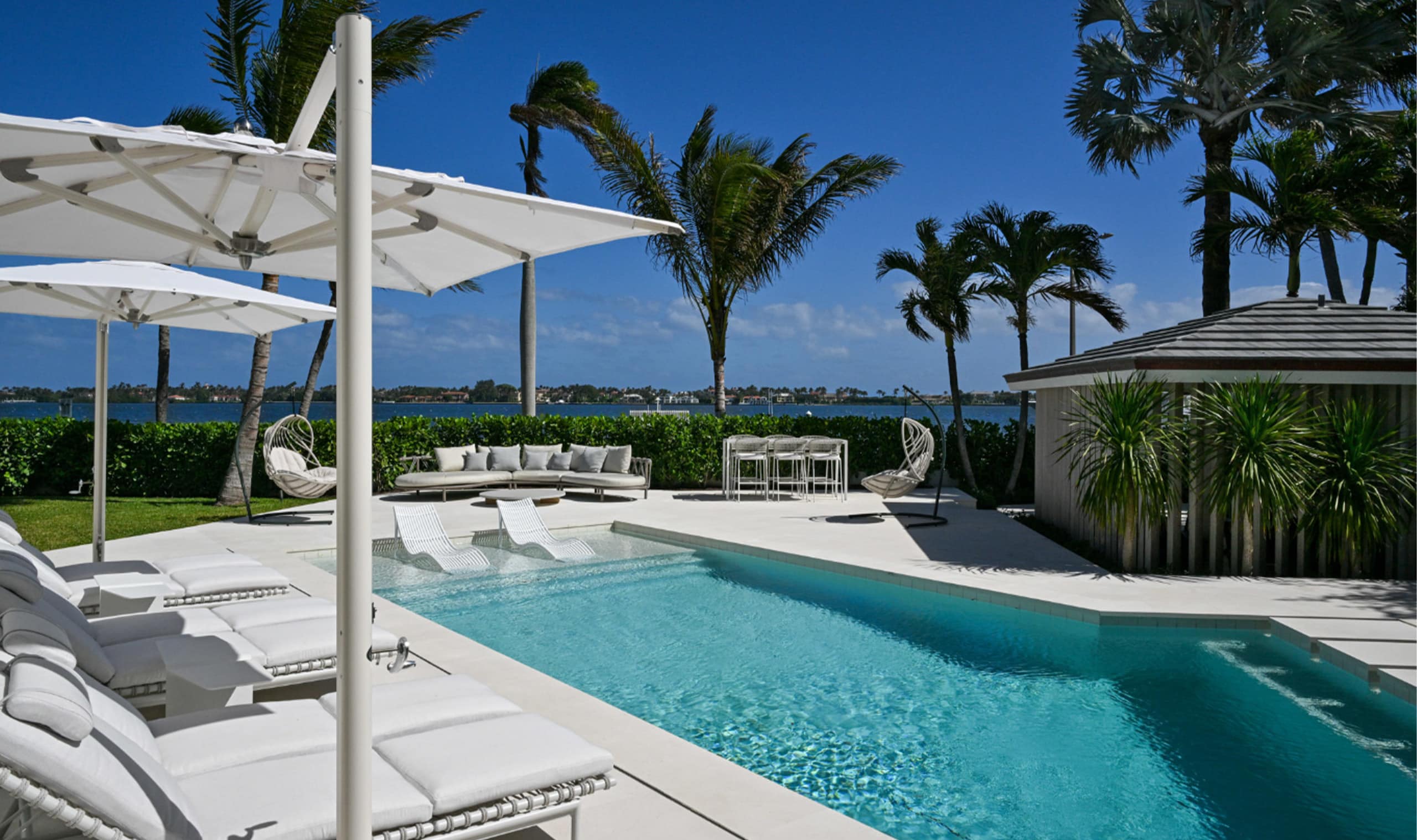 A serene waterfront retreat featuring a pristine pool, elegant white loungers, and lush palm trees against a bright blue sky