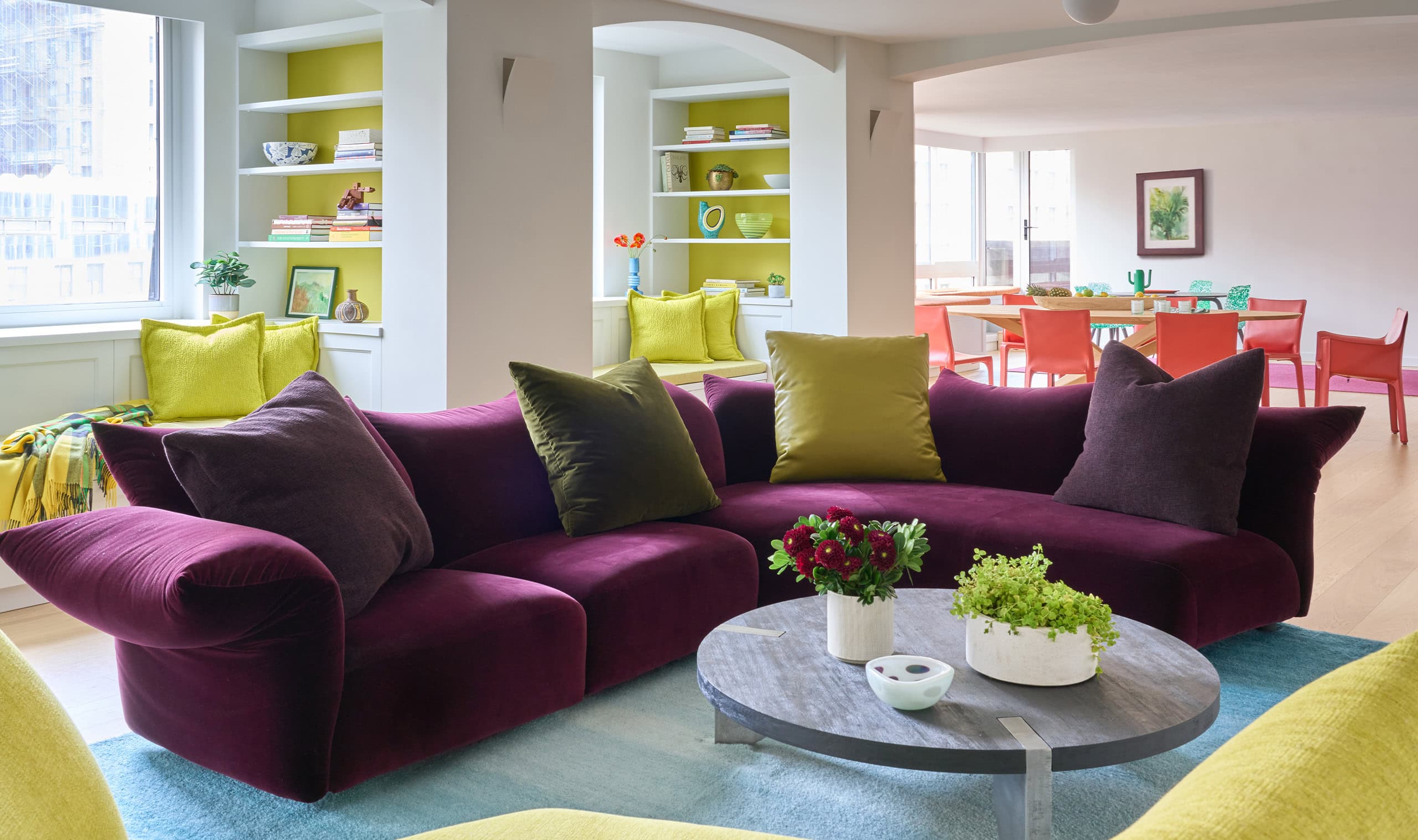 A vibrant living room featuring a plush purple sectional sofa, a round stone coffee table, and built-in bookshelves with bright accents