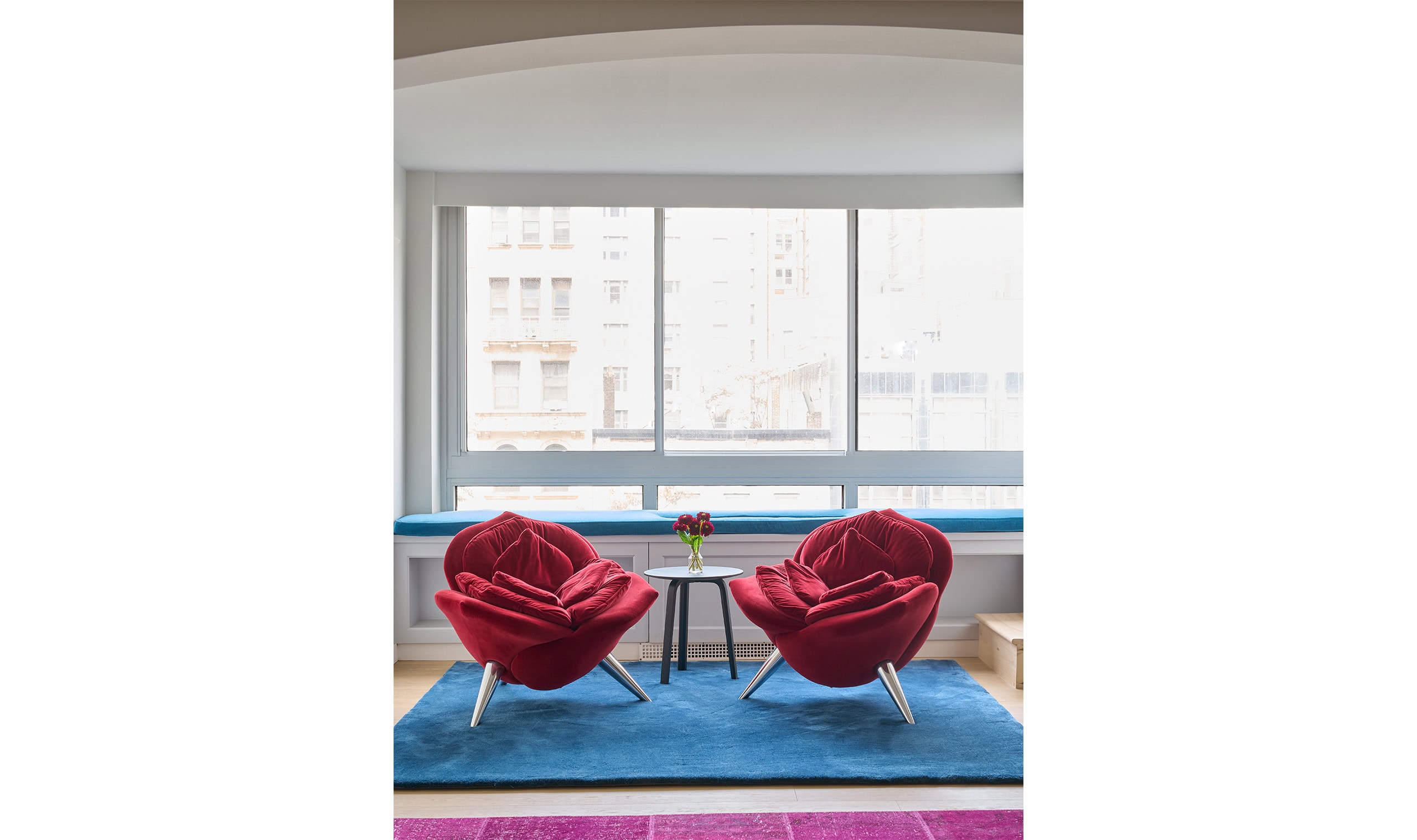 A contemporary seating area featuring two bold red accent chairs with a woven design, set on a blue rug in front of large windows
