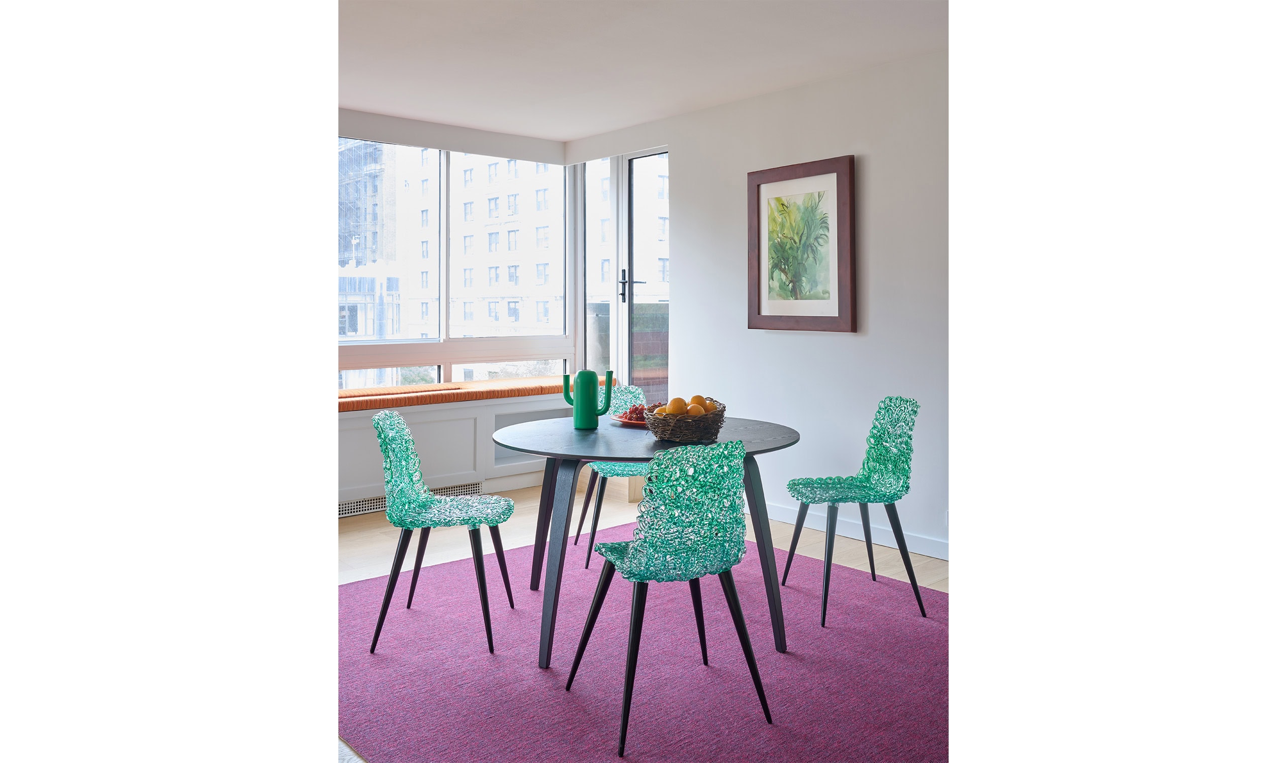 A modern dining space with a round black table, four vibrant turquoise chairs, and a magenta rug, set in a bright room with large windows