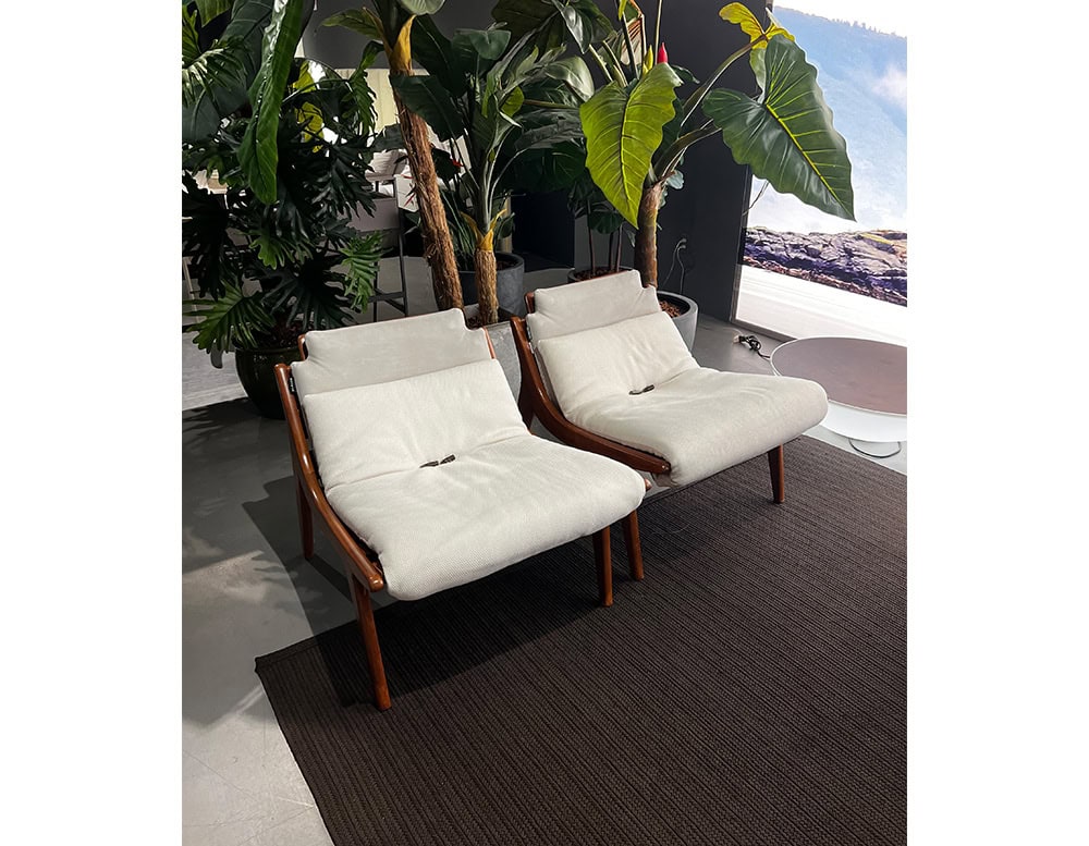 A stylish outdoor setting featuring the 10th Lerici Armchair with a sleek wooden frame plush light-colored cushions and lush greenery in the background