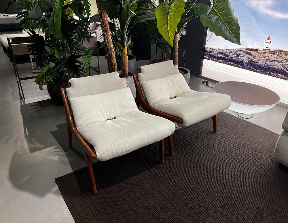 A stylish outdoor setting featuring the 10th Lerici Armchair with a sleek wooden frame plush light-colored cushions and lush greenery in the background