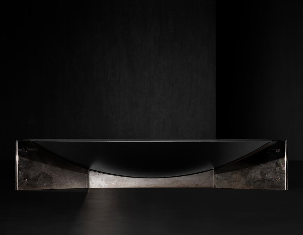 The Muse Coffee Table is a refined contrast of materials, featuring a deep black circular top resting on a cross-braced base of oxidized metal. The composition highlights elegant curvature and rich tones while maintaining a clean, minimalist profile.