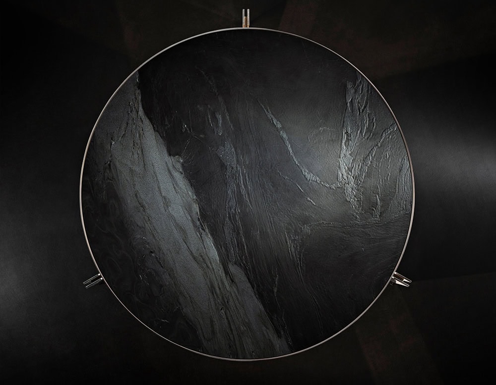 The Muse Coffee Table showcases a round, dark stone top with striking veining, bordered by a refined metal edge. Its sculptural silhouette and rich materiality evoke quiet strength and elegance in a minimal composition.