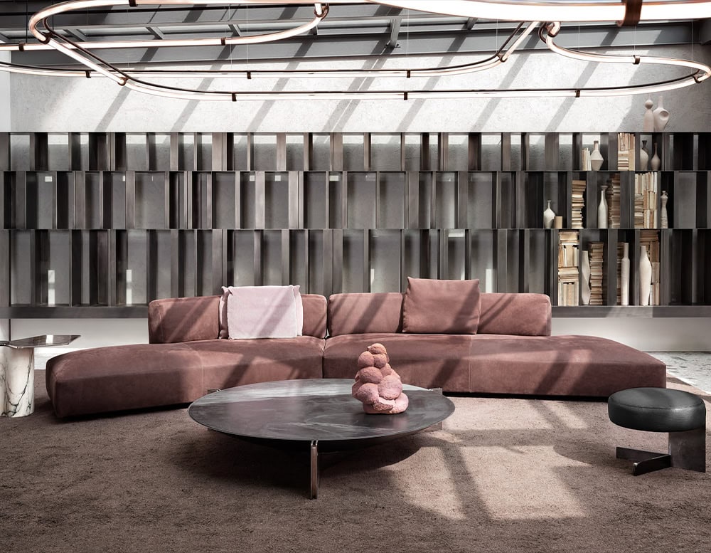 The Muse Coffee Table anchors the room with its dark stone top and steel base. Surrounded by rose-toned seating and shadowy shelving, the space blends rich textures and subdued hues into a warm, modern composition.