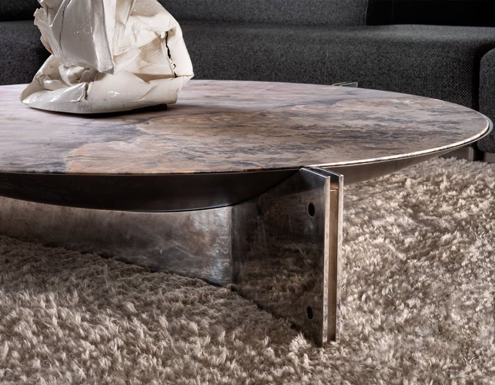 The Muse Coffee Table features a warm-toned stone top with rich brown and grey veining, set on a polished steel base. Paired with a dark charcoal sofa and a cream sculptural centerpiece, it rests atop a soft beige shag rug.