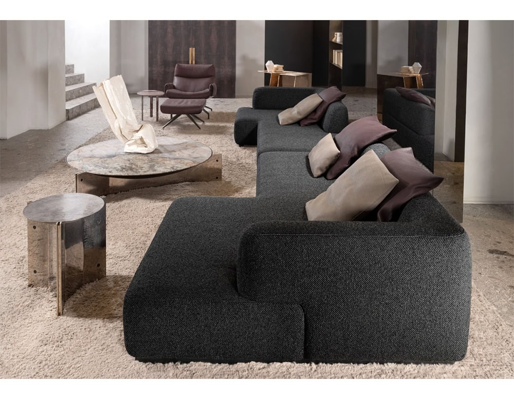 The coffee table features a beige and gray stone top with a polished metal base. It anchors a charcoal modular sofa layered with taupe and plum pillows, set against a textured cream rug for a warm and grounded look.