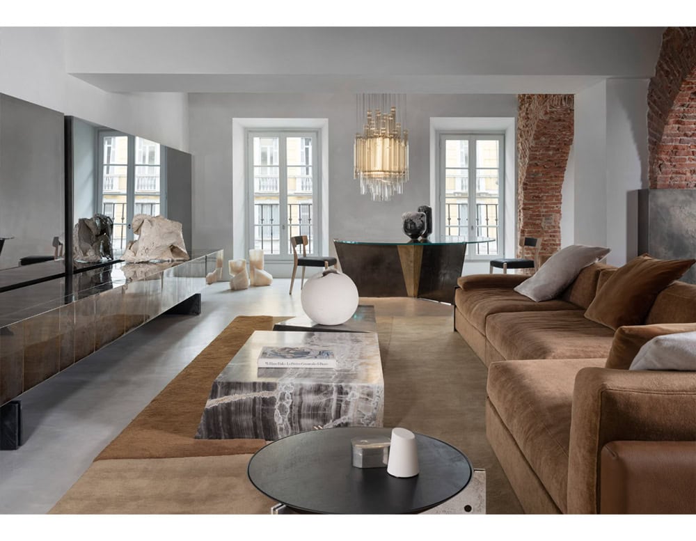 Hen Muse Coffee Table anchors the space with smoky gray and ivory veining, paired with a plush brown sofa and soft cream accents, all grounded by earthy neutral rugs and exposed brick columns.