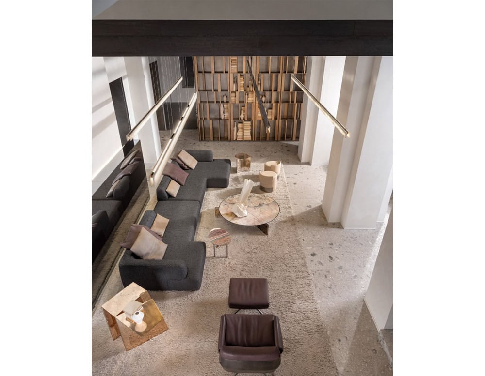 Hen Muse Coffee Table takes center stage with its soft marbled tones of beige and gray, surrounded by a deep charcoal sectional, mauve and cream pillows, and paired with sculptural beige stools and neutral textures throughout the room.