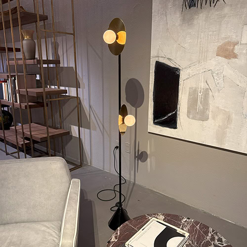 Ate Disc and Sphere Floor Light presents twin glowing orbs framed by golden discs on a sleek black stand. It stands beside a contemporary art wall and open brass shelving, offering a warm, sculptural accent to the modern setting.