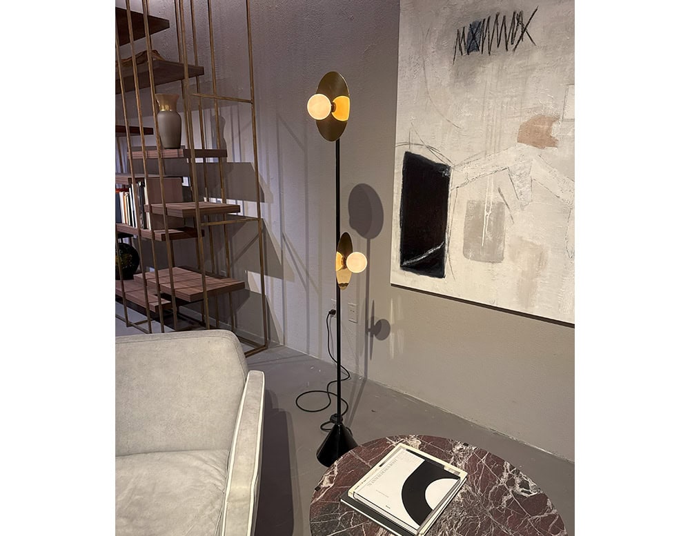 Ate Disc and Sphere Floor Light features twin globes on a slim black stand with golden reflectors, casting a warm glow. Its sculptural form adds elegance between a marble coffee table and modern shelving.