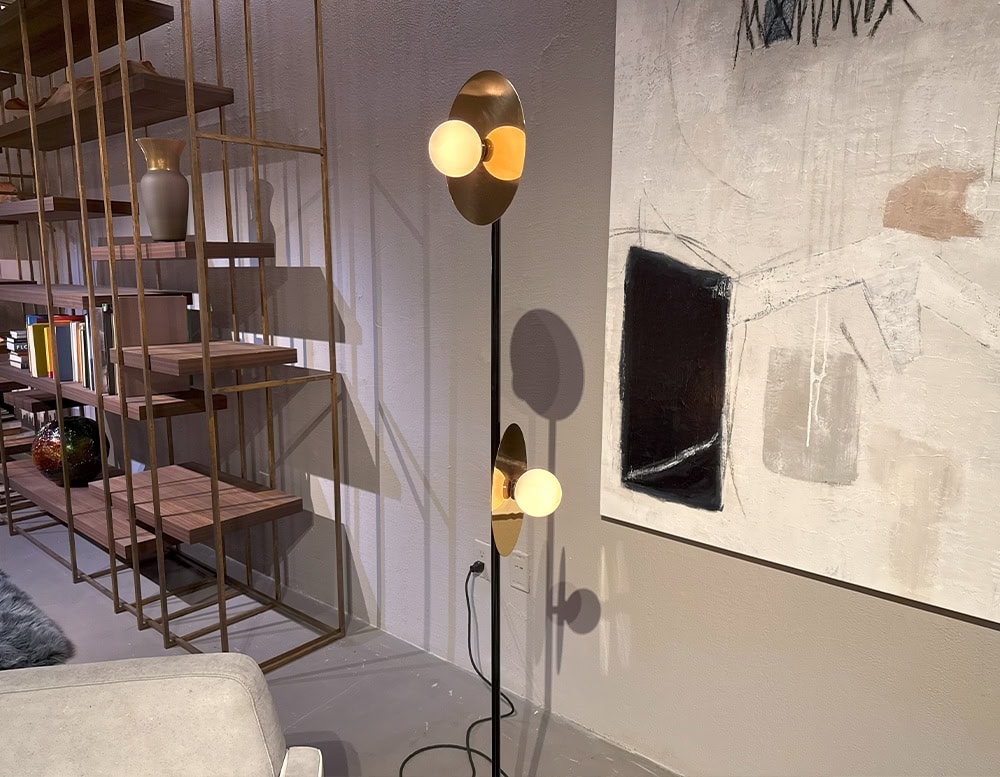 Ate Disc and Sphere Floor Light features dual glowing orbs set against brass discs along a slender black frame. It casts soft reflections across nearby art and open shelving, enriching the modern room with sculptural rhythm and warm light.