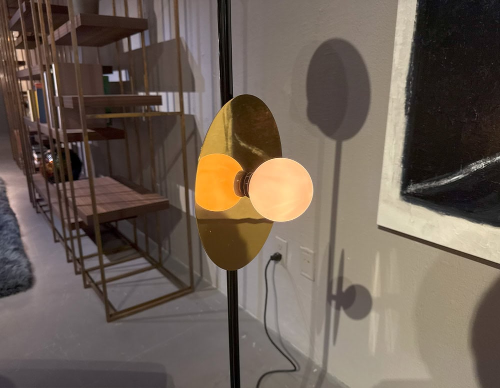 Ate Disc and Sphere Floor Light displays a glowing orb balanced in front of a reflective brass disc on a slender black pole. Nearby bookshelves and abstract art add dimension to the softly lit scene.