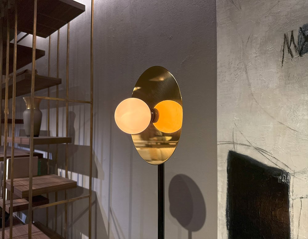 Ate Disc and Sphere Floor Light showcases a glowing opal sphere set against a polished brass disc, creating a warm halo effect. Set in a moody interior with open shelving and textured wall art, it adds sculptural intrigue and ambient light.