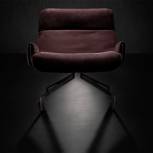 The Hen Club 57 Armchair features plush burgundy upholstery with a deep seat and sleek, angled arms. Its metal swivel base adds a modern edge, set against a dark, moody background for dramatic contrast.