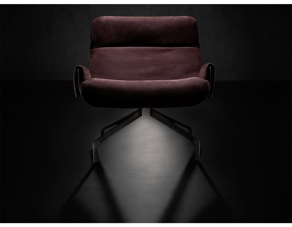 The Hen Club 57 Armchair showcases deep burgundy upholstery with smooth contours and slim armrests. Its sculptural metal base provides contrast, highlighted by focused lighting against a dark backdrop.