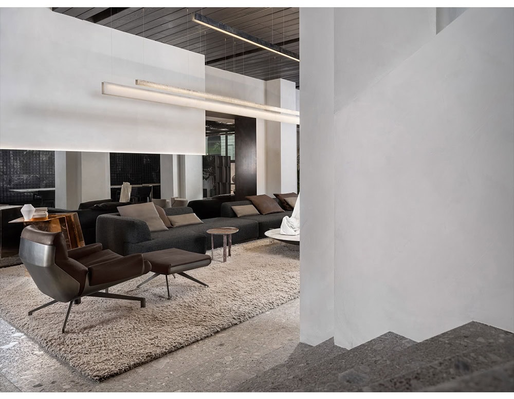 The Hen Club 57 Armchair and ottoman are shown in a lounge setting, with a brown leather finish and angular metal base. They rest on a soft beige rug, surrounded by dark sofas and minimalist architectural elements.