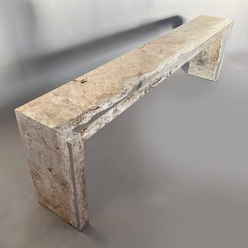 The Rapolano Console Table features a solid travertine form with raw, textured edges that highlight its natural stone composition. The design is minimalist, with clean lines and a sculptural presence.