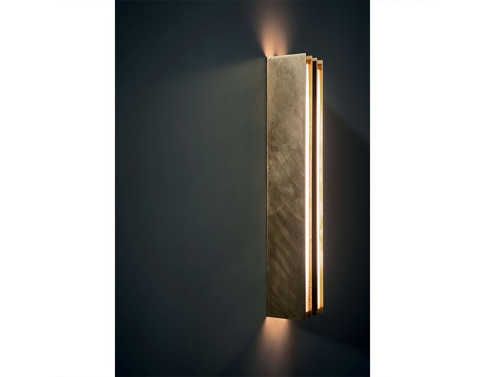 Bax Blade Wall Light showcases a bold vertical form in brushed metal, emitting a soft glow from its recessed edges. Mounted on a deep-toned wall, the piece casts delicate light both upward and downward, accentuating its sculptural presence.