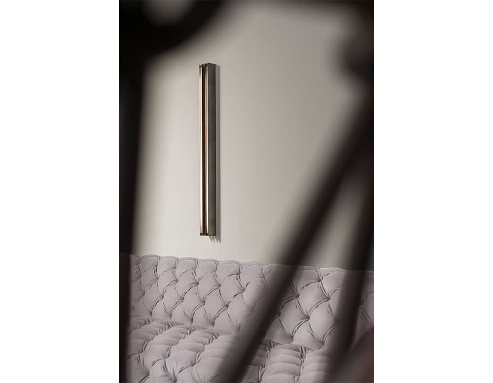 Bax Blade Wall Light features a sleek vertical bar mounted on a pale wall, casting soft illumination. Set above a tufted, plush sofa in a muted hue, the minimal light enhances the cozy and refined ambiance of the space.