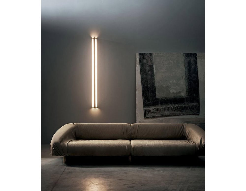 Bax Blade Wall Light is a pair of vertical glowing bars casting warm light along a dark wall. Positioned behind a deep-toned, plush sofa and beside a bold abstract painting, the light adds drama to the subdued, moody setting.