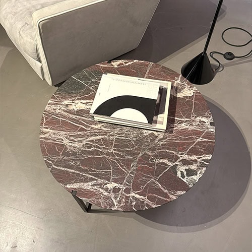 Bax Icaro Coffee Table stands out with its rich red marble top and white veining, supported by a slender black frame. A few books are placed on top beside a modern lamp and neutral-toned seating in a calm, softly lit setting.