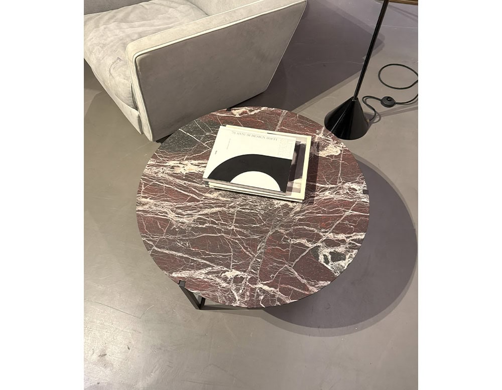 Bax Icaro Coffee Table features a round red marble top with dramatic veining and a minimalist black metal base. A few books rest on the surface beside a modern floor lamp and pale seating, creating a quiet reading corner.