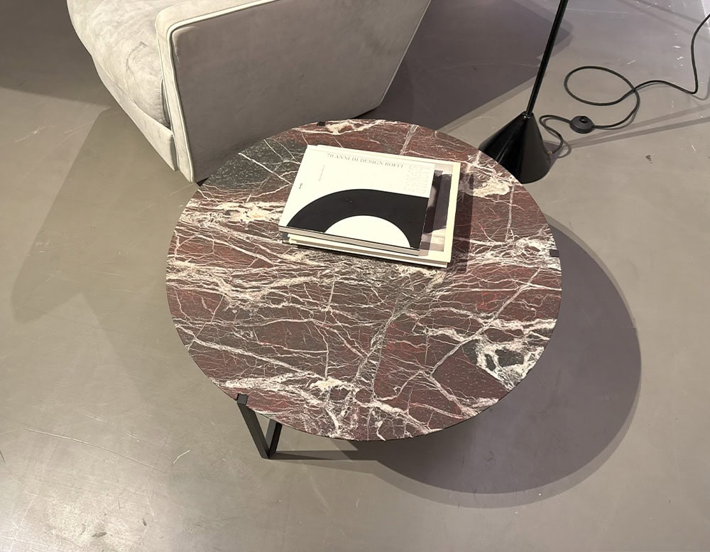 Bax Icaro Coffee Table has a round red marble top with white veins and rests on a sleek black metal base. A stack of books is placed on it while a floor lamp and upholstered seating subtly frame the cozy setting.