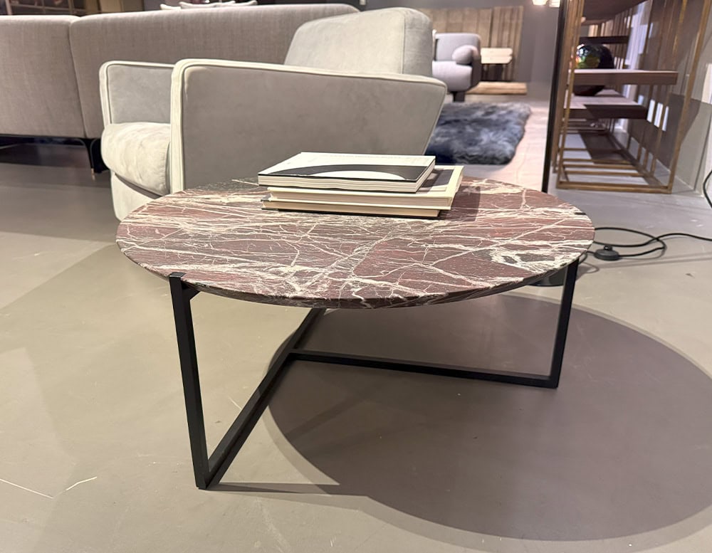 Bax Icaro Coffee Table features a round red marble top with fine white veining, supported by a black angular metal base. It sits on a smooth floor, surrounded by neutral sofas and layered rugs in a cozy modern lounge setting.