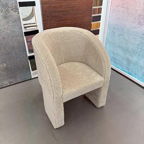 Bax Lazybones Armchair is upholstered in a light beige fabric with a soft nubby texture. The setting includes multicolored material panels and neutral floors that subtly frame the armchair’s clean rounded shape.