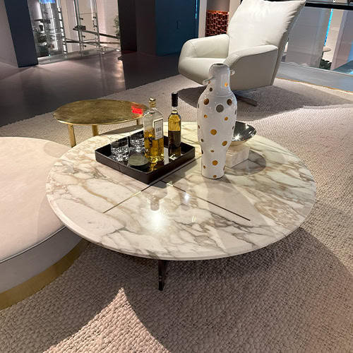 Bax Place Coffee Table featuring a smooth circular marble top with natural veining and slim dark legs. Styled with bottles, a polka dot vase, and surrounded by soft seating, it anchors a cozy and refined lounge space.