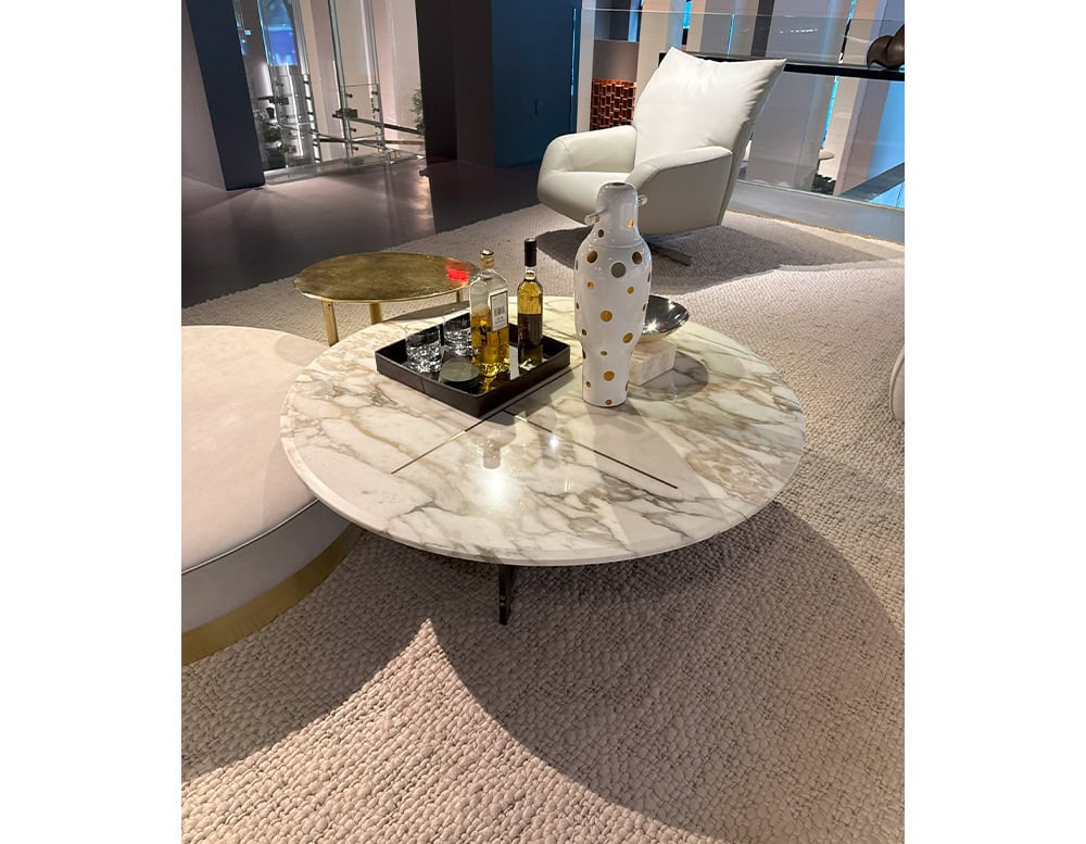 Bax Place Coffee Table with a light veined marble top and slender dark legs. Styled with bottles, glassware, and a tall dotted vase, the table sits among plush seating and textured flooring in a calm and softly lit lounge arrangement.