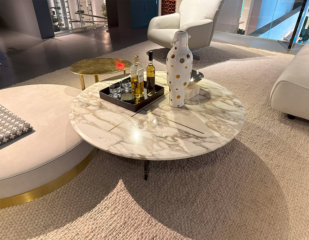 Bax Place Coffee Table with a round marble top veined in soft beige tones and supported by slim metal legs. It anchors a relaxed arrangement with a sculptural white vase, drink tray, and soft seating in a muted, plush setting.